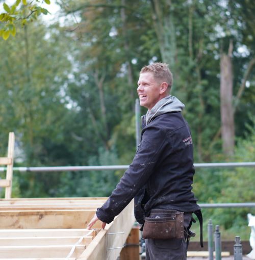 Voorman van Van Wiggen Bouw aan het werk op de bouwplaats, lachend tijdens het plaatsen van houten constructiedelen