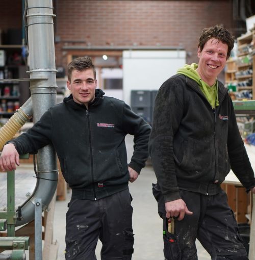 Een voorman en zijn maat bij van Van Wiggen Bouw in de werkplaats, lachend tijdens het voorbereiden van houtwerk