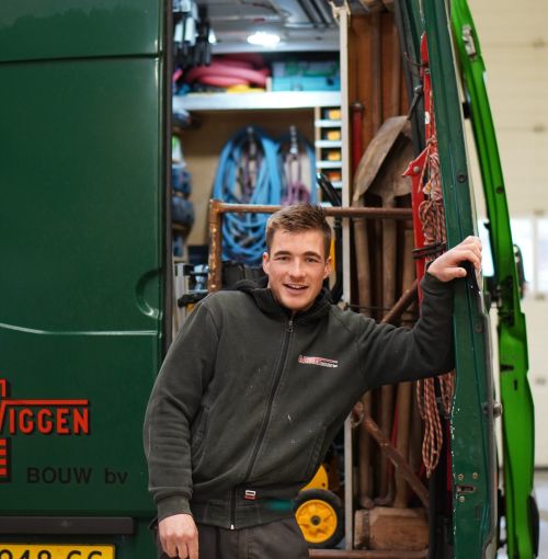 Timmerman van Van Wiggen Bouw leunend tegen de werkbus, klaar voor vertrek naar de bouwplaats