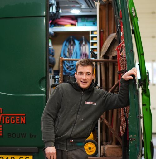 Timmerman van Van Wiggen Bouw leunend tegen de werkbus, klaar voor vertrek naar de bouwplaats